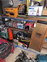 Wide view of shelving with various boxed auto parts, tools, cords, an industrial light, and a Craftsman auto creeper leaning against shelves.