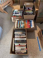 Four cardboard boxes filled with hardcover books stacked upright and lying down. The tops of the books show a variety of titles and authors in fiction and nonfiction genres.