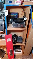 Photo showing wooden shelving with Bearcat 350A Scanner in box on top shelf, additional Uniden scanner on top shelf, Simpson 260 multimeter and black power supply on middle shelf, and cassette recorder on bottom shelf.