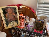 Wide view of collection on wooden surface showing boxed Cabbage Patch Kids Preemie doll, other dressed dolls, and plush toys including teddy bear and sheep, plus Rawlings mini mitt in box.