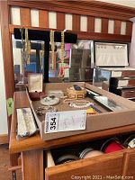 Photo showing a black jewelry display stand with multiple gold-tone chain necklaces and bracelets. Several pieces of costume jewelry are spread on the wooden desk surface including a pearl necklace and a wooden box.