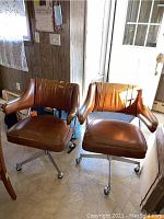 Two brown mid-century modern swivel chairs with chrome bases and casters side by side in a room near a door. The chairs have padded low backs and armrests with vertical stitching detail.