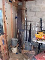 Corner view of saws, wooden crates, metal pails, hand axe, spool of rope, small lanterns, and wood beams.