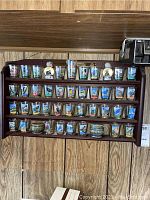 Photo of wooden shelf mounted on wall holding 33 different clear glass shot glasses with colorful songbird images and names displayed in 5 rows.