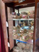 Wooden display cabinet with three glass shelves holding a collection of bone china tea cups and saucers.