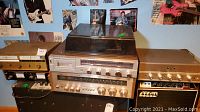 Three stacked audio components including gold-faced tuner, silver-faced turntable with dust cover, and gold-faced amplifier on a black table, posters in background