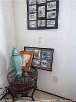 Overview showing round glass side table with rattan base, celadon vase on top, two framed artworks leaning behind.