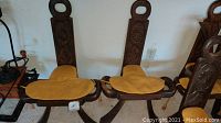 Two carved wooden saddle chairs with yellow seat pads, side view, showing the saddle shape and carved backs with circular handles.