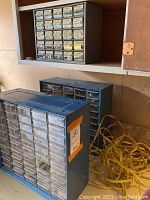 Three blue metal storage bins on shelves. Multiple small clear plastic drawers for organizing nuts, bolts, and small hardware. Coiled yellow strap next to them.