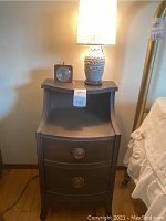 Full view of the vintage wooden bedside table with two drawers, Gilbert wind-up clock and Asian style lamp on top