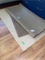 Two stacked rolled area rugs, beige underneath and brown on top, clearly showing their color and plush texture.