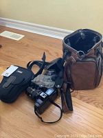 Leather Fotima camera bag next to various camera accessories including Minolta camera, lens, and filter parts.