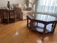 Coffee and end table set next to floral upholstered chair in a room with wood flooring and large windows with sheer curtains.