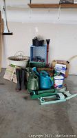 Wide view of garden supplies on the floor showing planters, watering cans, boots, garden tools, gas can and hose.