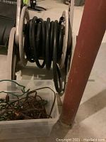 Plastic hose caddy on wheels with coiled black hose, pictured in basement next to red metal pipe and plastic bin.