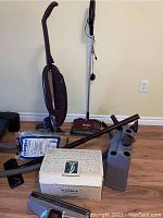 Kirby Performance 5G vacuum and Shark 2-speed vacuum, with multiple accessories including hoses, attachments, vacuum bags, and shampoo canisters arranged on a wooden floor.