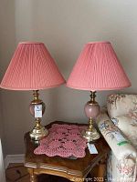 Pair of table lamps with pink ceramic bases, brass-look details, pink pleated shades, and matching pink crocheted doily on wooden side table.
