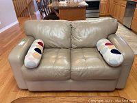 Front view of beige leather and faux leather love seat with two multicolored cylindrical throw pillows on a hardwood floor in a kitchen/living room area.