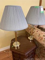 Pair of matching table lamps on a wooden side table against a beige wall, showing glass and gold-coloured pillar bases with gray pleated fabric shades.