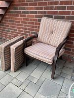 Two brown metal lawn chairs with matching striped cushions, placed on a brick tile patio against a brick wall. Cushions are separate and thick.
