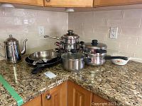Countertop display of stainless steel coffee pot, two frying pans (one non-stick), stacked pots with lids, and Corning casserole dish with handle.