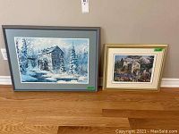 Two framed prints by Keirstead placed on the floor against a wall; left is larger winter mill scene, right is smaller 'Brenda's Garden'.