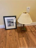 Framed signed outdoor rink print with black frame and white matting next to a 17-inch candlestick-style lamp with brass-colored base and beige pleated lamp shade on a hardwood floor.