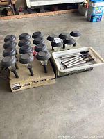 Photo showing 12 solar garden stake lights with round black tops on a cardboard base and 5 solar garden stake lights with tiered black tops next to white tray with stakes.