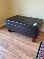 Front-side view of the brown vegan leather storage bench showing tufted top surface and wooden legs on a hardwood floor.