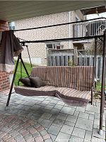 Front view of the large outdoor metal glider swing with brown striped cushions and two pillows. Metal canopy frame visible without fabric canopy attached.