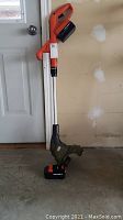 Full view of the weed trimmer standing upright showing the orange and black body, silver shaft, and battery attached at bottom.