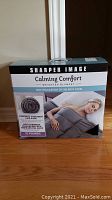 Front of the original box showing brand Sharper Image, product name Calming Comfort weighted blanket, with details about relaxation benefits and weight of 15 pounds.