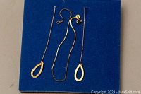 Three gold jewelry items arranged on a blue background: a thin 7-inch gold chain bracelet and two teardrop-shaped gold earrings, one smooth and one textured.