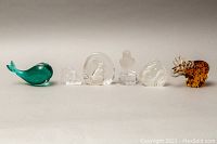 Full view of six small glass figurines lined up showing variety of animals and abstract pieces including green whale, clear glass owl and moose.