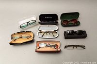 All eight pairs of glasses with cases except one pair without case, arranged flat on a light surface. Various styles and colors.