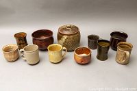 Photo of the whole lot showing ten assorted pottery pieces including mugs, chip bowls, a lidded jar, and planter pots in earthy glaze tones.