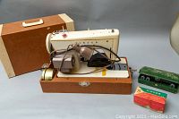 Wide view of vintage Singer sewing machine mounted in brown hard case with attached lamp and brown foot pedal, also showing white carrying case behind.