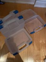Four translucent plastic craft totes with blue latches arranged on floor, showing empty interior and open lid.