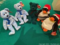 Five Ty Christmas Beanie Babies including two blue bears with snowflake patterns, a green bear, and two penguins wearing Santa hats on a green surface.
