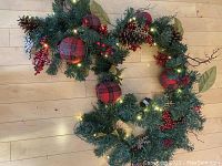 Full view of the green artificial Christmas garland decorated with red berries, pine cones, plaid fabric balls, large green leaves, and white lights.