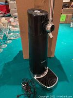 Fizzics drink machine standing upright with attached power cord, positioned in front of a cardboard box on a green surface.