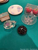 Photo showing all lot items together on green surface including two flower frogs, rabbit tea light holder, vintage glass box, tomato salt and pepper shakers in packaging, and China plate.