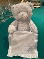 Light gray Gund Peekaboo Bear holding a white blanket with satin trim and embroidered logo, sitting upright on a teal surface.
