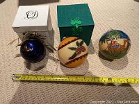 Photo showing three large glass tree ornaments laid on carpet next to measuring tape indicating each is about 4 inches in diameter. One is deep blue with gold writing and housed in white box, the other two are painted glass spheres with different themes, one in green box and one without a box.