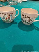 Two large ceramic mugs with colorful Maw and Paw cartoon designs and text sitting on blue surface.