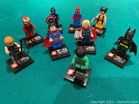 Wide shot showing all eight superhero mini brick figures on green background. Figures include Iron Man, Captain America, Spider-Man, Wolverine, Superman, Thor, Batman, and Hulk each on their own black stand.
