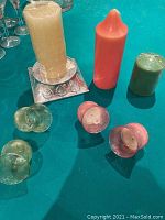 Photo showing three pillar candles of beige, coral, and green colors along with four small green glass candle holders and a large silver candle stand on a teal surface.