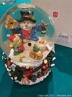 Clear glass snow globe featuring a snowman holding a lantern and a child with a red hat inside a snowy winter scene, base decorated with holly and berries.