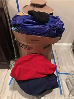 Cardboard box containing multiple blue folded bags along with red and navy blue hats on top and in front of the box.