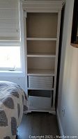 Full view of tall white wooden bookcase with two wicker baskets on lower shelves beside a bed and window.